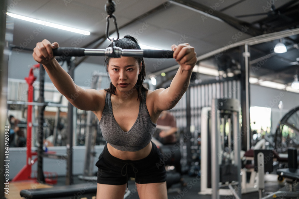 Fototapeta premium Determined young woman performing standing cable press in a modern gym. Healthy lifestyle and wellness concept.