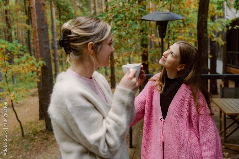 Fototapeta premium Two friends share a joyful moment in a serene forest setting, sipping warm drinks. Surrounded by trees with autumn leaves, they enjoy each other's company on a chilly day.