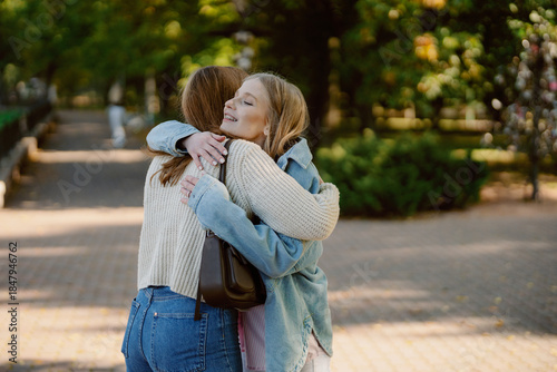 Two friends share a cheerful hug in a beautiful park. Surrounded by lush greenery, their smiles brighten the warm afternoon as they enjoy each other's company and friendship.