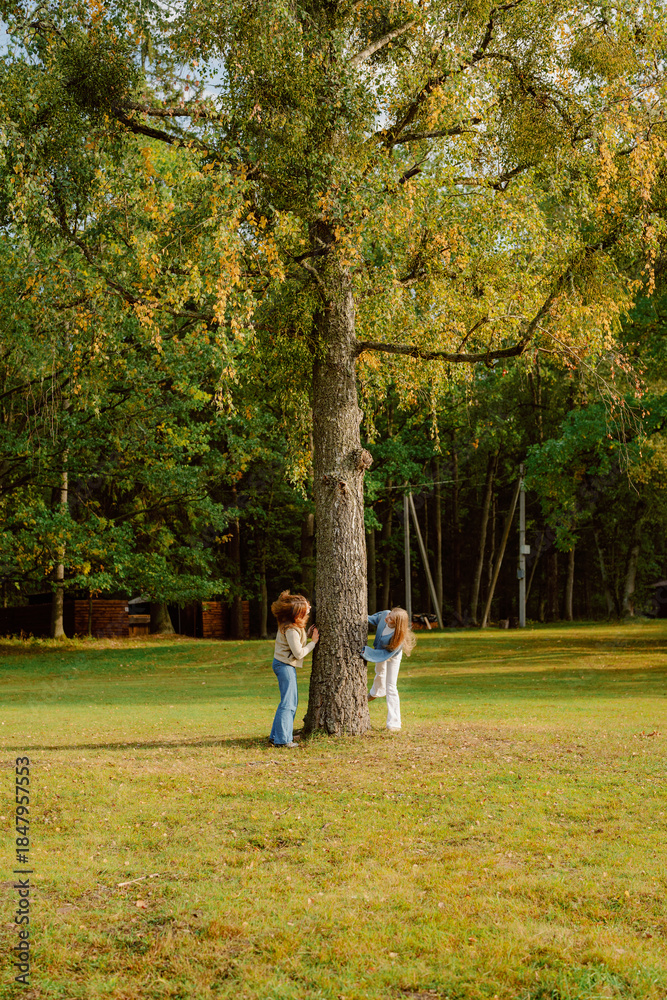 Fototapeta premium Two friends enjoy a fun afternoon in a lush green park, playfully hiding behind a tall, sturdy tree. The sunlight filters through the leaves, creating a warm, inviting atmosphere.