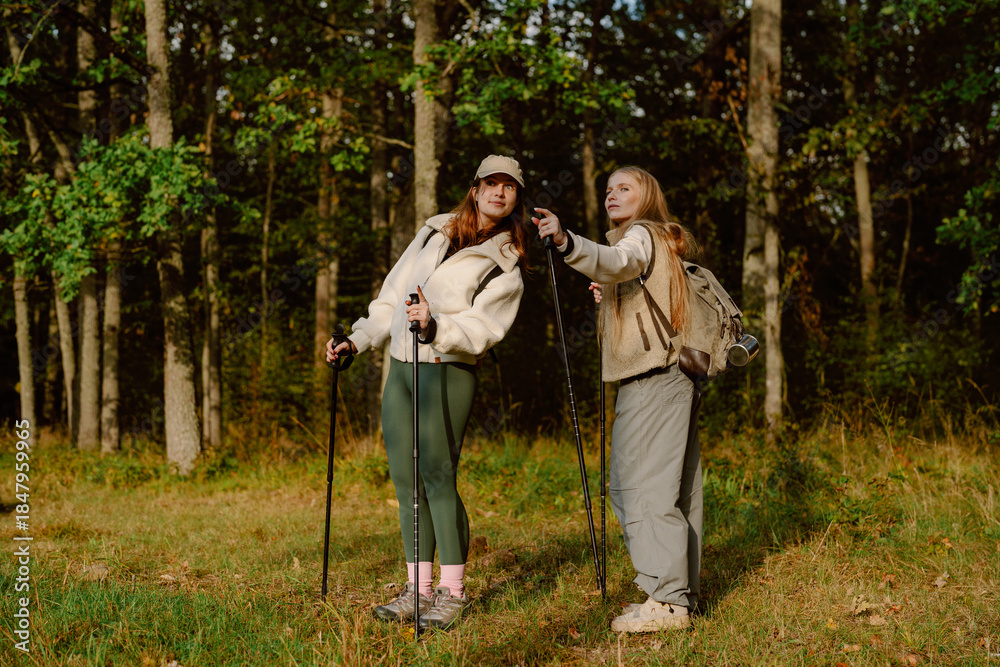 Obraz premium Two young women enjoy a lively hike through a forested area. They discuss the scenery while using trekking poles, surrounded by lush greenery on a beautiful day.