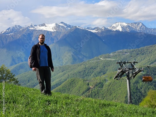 Man standing on a hill with a green background with mountain and cable way. He is wearing a blue shirt and black jacket