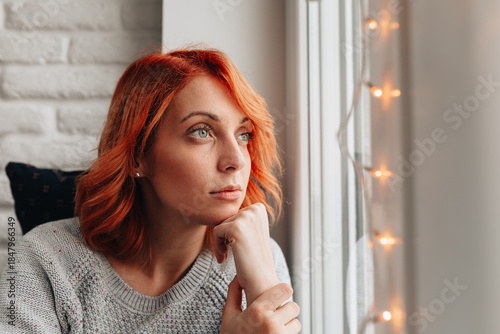 Woman with orange hair looks out the window while resting her chin on her hand in a cozy indoor space during daytime