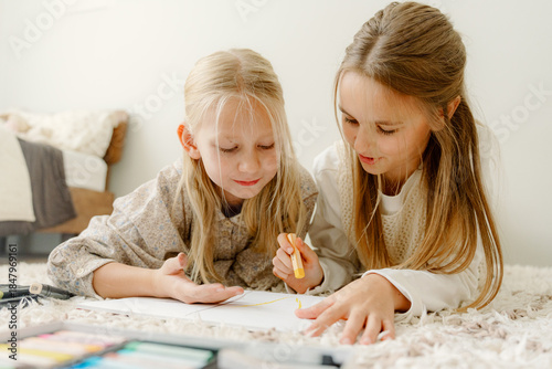 Photography Two young girls lay on a soft rug, happily focused on creating drawings with colorful markers