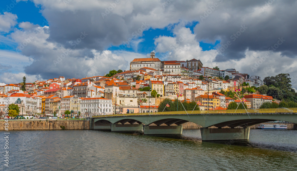 Obraz premium Scenic view of Coimbra city in Portugal with the University on the hill, Mondego River, and Santa Clara bridge under a cloudy blue summer sky.