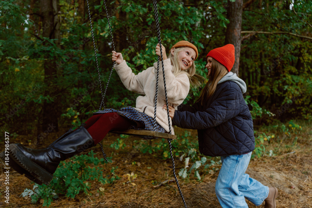 Obraz premium Two friends have a lively moment in a sunlit forest. One friend swings joyfully while the other pushes, both wearing cozy autumn outfits. Laughter and fun fill the air.
