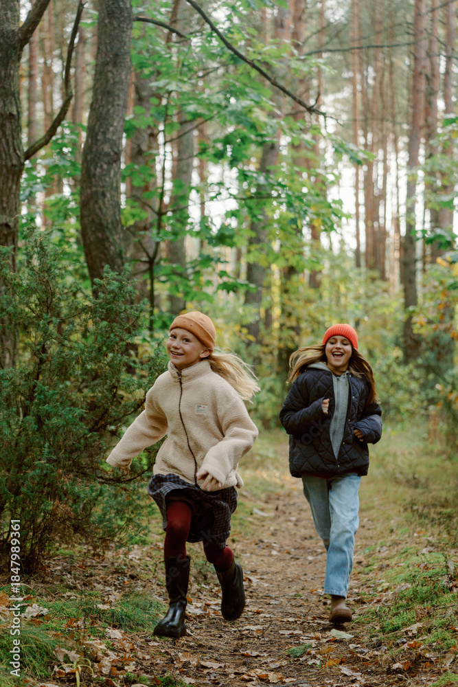 Fototapeta premium Two young girls are running excitedly down a narrow path in a vibrant forest. The trees are lush with green leaves, and the sun is shining through, creating a cheerful atmosphere.