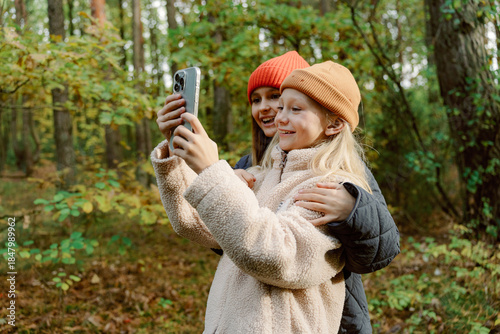 Two girls stand together in a lively forest, smiling as they take a selfie. Their cozy outfits and bright hats contrast beautifully with the fall foliage surrounding them.