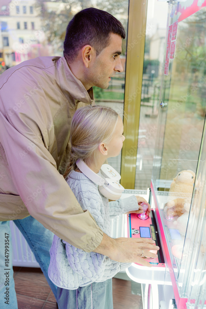 Obraz premium A father and his young daughter enjoy a lively afternoon at an arcade, eagerly trying to win a stuffed toy from the claw machine. Their expressions show excitement and joy.