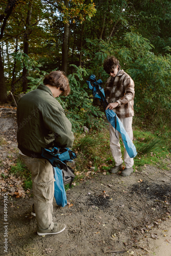 Naklejka premium Two friends are preparing to set up a tent in a serene forest. With trees adorned in vibrant autumn leaves, the scene is peaceful and full of nature's beauty.