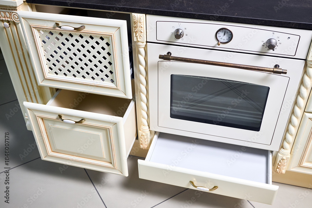 Fototapeta premium Interior of modern kitchen in classic style with golden elements in beige colors with built-in appliances. Drawer for plates or dishes shelf under the countertop with lattice front panel made of wood.
