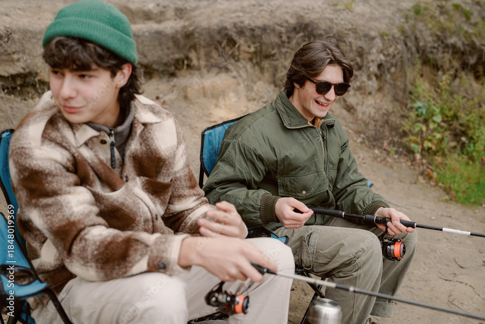 Fototapeta premium Two young men sit beside a river, fishing rods in hand, surrounded by nature. One smiles while the other looks out at the water, enjoying a relaxed day outdoors.