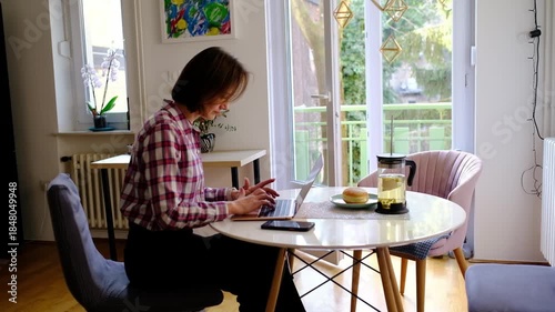 Smiling adult woman in works from home in sunny kitchen, typing on laptop, enjoying work-life balance, singing joyfully, feeling relaxed and inspired by remote job freedom.