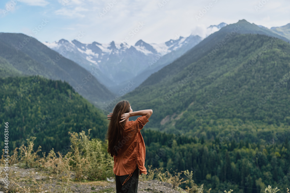 Fototapeta premium A lone traveler stands on a hillside, gazing at snow capped mountains and a vast valley, soaking in the tranquil alpine landscape and dense forest, creating a moment of awe and quiet reflection.
