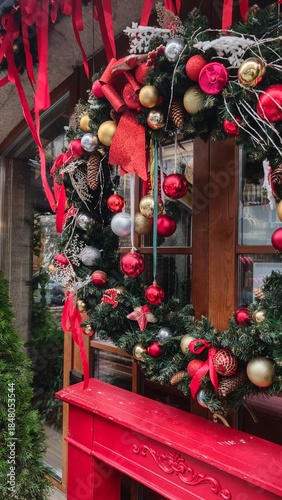Colorful Christmas wreath with red, gold and silver ornaments hangs on a wooden door with festive ribbons. Concept of traditional holiday spirit, warm welcome and seasonal decoration