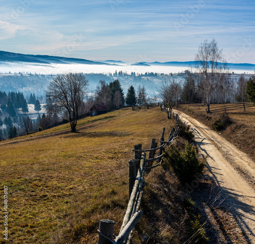 Countryside Mountain Road with Wooden Fence in Rural Landscape. Sunshine in the late autumn mountain landscape with a fence and dirt road above misty valleys, Harghita County, Livezi village, Romania.