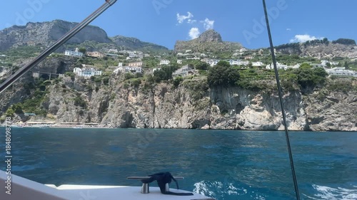 Cruise summer vacation on luxury yacht along famous Amalfi coastline, wake foam on water surface from riding speed boat, panoramic view of rock hills with greenery and colourful scattered houses