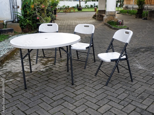white circular folding table and three matching plastic chairs arranged on a grey brick-paved courtyard. The background features tropical greenery and a colonial-style building.
