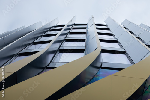 Modern building with curved metallic facade and large windows viewed from below against cloudy sky with reflections on glass surfaces and architectural details
