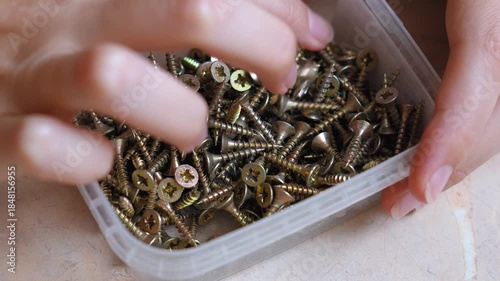 Close up view of female hands holding a transparent plastic container and sorting through a pile of small metal screws, searching for the right size for a home improvement or diy project