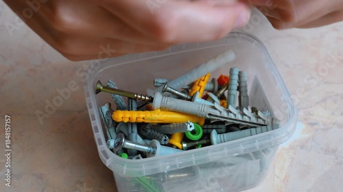 Woman's hands searching and sorting through a plastic container filled with a variety of colorful wall plugs, dowels, and metal screws for home improvement and diy construction projects