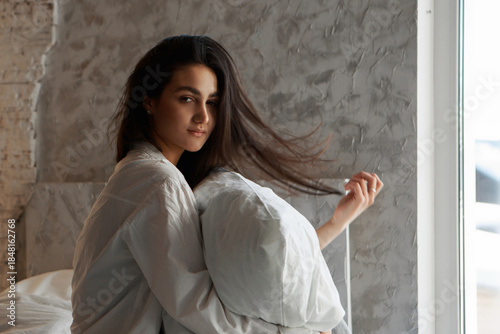 A woman with black hair wakes up in the morning in a white blanket and catches the rays of the sun. The portrait is creative, with brown eyes. High quality photo