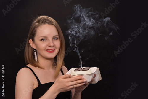 A woman in a black dress blows out a candle on a bento cake. Smoke on a black background. A girl's birthday, happiness, a gift. High quality photo