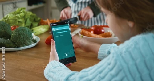 Young girl uses an AI chat app with voice input on a smartphone to ask for a salad recipe and shows it to her father while cooking together in the kitchen. Everyday AI technology in family life.