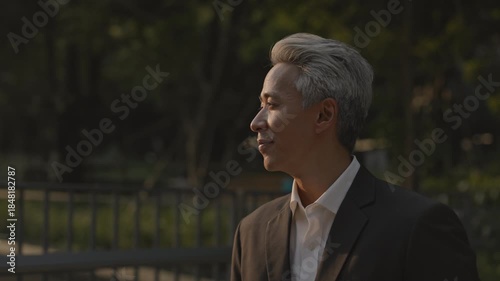 Portrait of mature Asian businessman looks around and walks along footbridge in sunset downtown. Cheerful Japanese entrepreneur in formal attire observes cityscape in town
