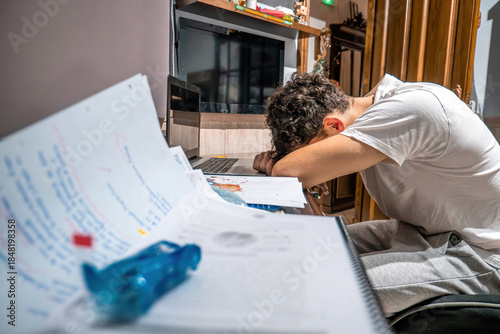 Young man feeling exhausted and overwhelmed, sleeping with head on his desk surrounded by books and laptop after intense studying