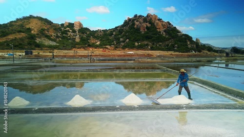 Asian worker harvests sea salt crystals in solar evaporation ponds with rocky hills in background. Traditional mineral production in coastal countryside of Vietnam.