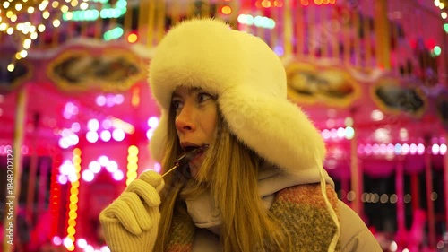 Cheerful young woman in a fur hat enjoying a rooster-shaped lollipop while visiting a festive christmas market at night, with the colorful blurred lights of a carousel in the background