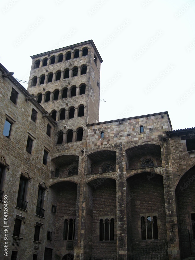 Fototapeta premium Exterior view of a tall, historic medieval stone tower with multiple rows of arched windows adjacent to an ancient palace complex in the Gothic Quarter of a city