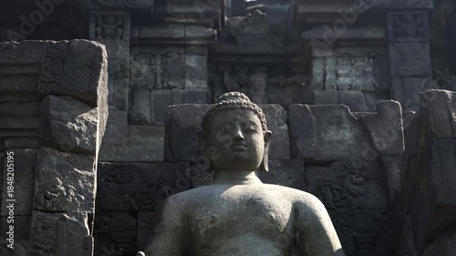 Statues of Buddha sitting in Vara mudra inside wall niches illuminated by sun. Beautiful decorative details of Borobudur Mahayana Buddhist temple, Magelang, Central Java, Indonesia. Camera zooms out.