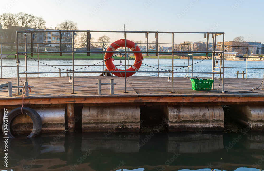 Fototapeta premium Floating pier with metal railings, a bright lifebuoy ring, and mooring hardware above still water. Simple geometric composition for safety, rescue, waterfront infrastructure, and maritime travel conce