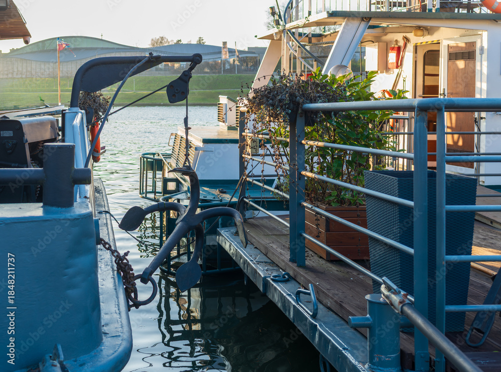 Fototapeta premium Close view of dock railings, anchor shape, and moored boats on calm water during golden hour. Industrial maritime details meet quiet waterfront mood, useful for travel, harbor, and nautical design bac