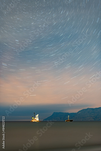 Star trails over shipwreck and anchored cargo ship at night