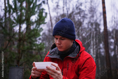 A young man eating instant noodles in nature. Lunch during a hike in the forest, fast food at a picnic