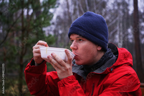 A young man eating instant noodles in nature. Lunch during a hike in the forest, fast food at a picnic