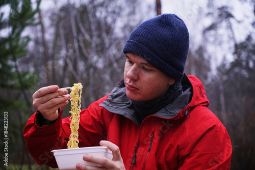 A young man eating instant noodles in nature. Lunch during a hike in the forest, fast food at a picnic