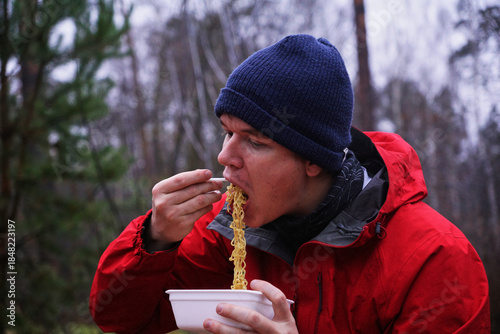 A young man eating instant noodles in nature. Lunch during a hike in the forest, fast food at a picnic