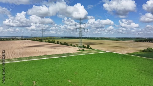 Slow side drone flight around a high-voltage pylon with the transmission line crossing the landscape, surrounded by cultivated and harvested fields under a blue sky with clouds.