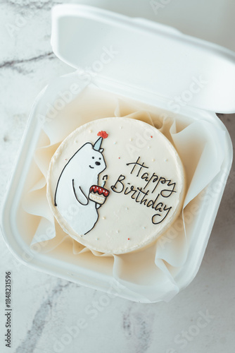 Cute bento cake with funny white bear holding a festive cake with a candle drawing. Birthday cake in a white gift box on the white marble background