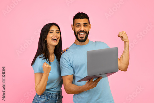 Two young adults express joy and excitement while holding a laptop in front of a bright pink background. They appear to have achieved something noteworthy together.