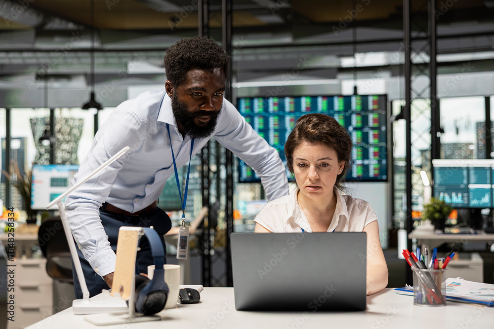 Fototapeta premium Multiethnic coworkers discussing projects at desk. Black male executive and female employee using laptop on table to analyze reports, track statistics and plan corporate growth strategies.