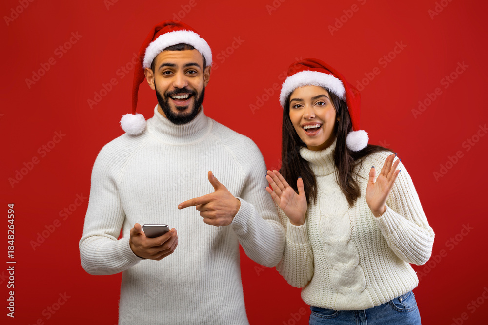 Fototapeta premium A cheerful Arab couple wearing Santa hats is happily surprised while receiving a Christmas offer on a smartphone. They are posing together in front of a red background.