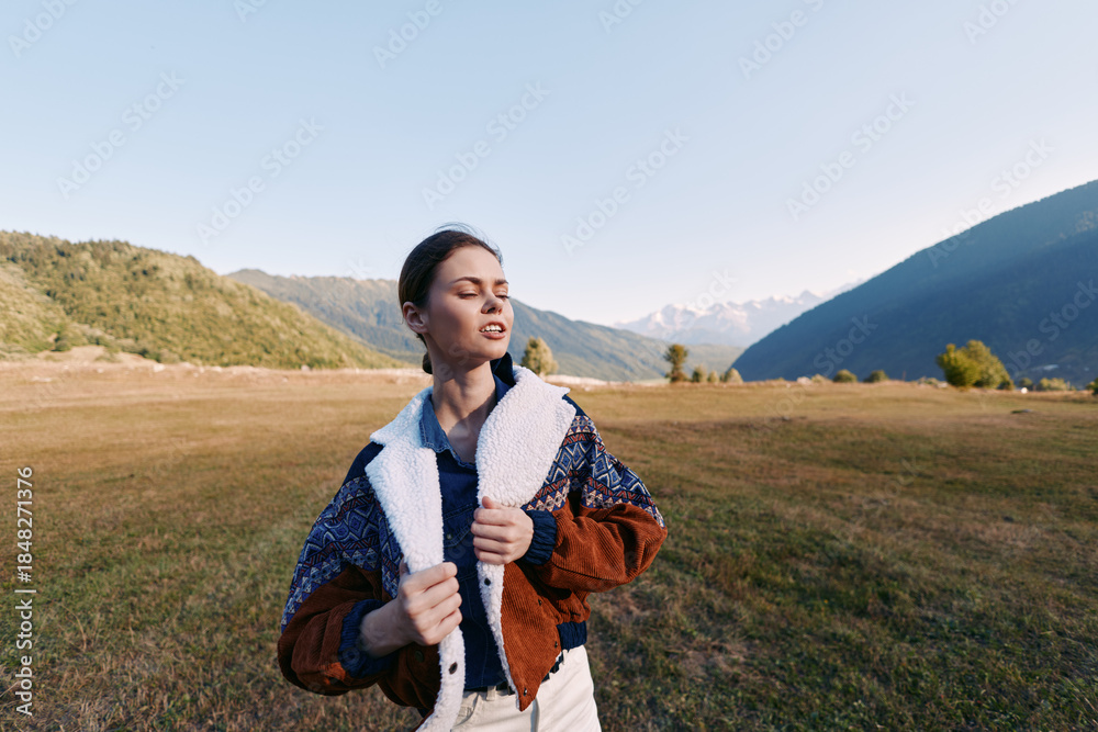 Fototapeta premium Woman portrait in nature standing on a wide meadow with mountains in the background, wearing a warm jacket during travel outdoors. Young person enjoying sunlight, scenic landscape and calm horizon.