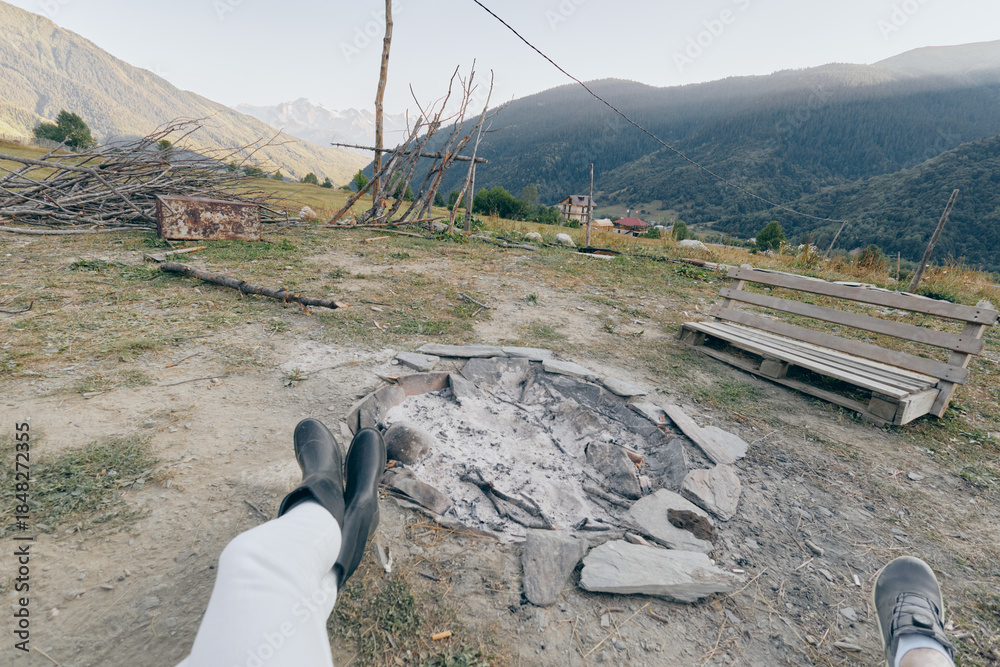 Fototapeta premium Campfire mountains boots bench firepit hiking outdoors relaxation. Two people resting with boots stretched toward a stone firepit and wooden bench, panoramic mountain valley view at campsite.