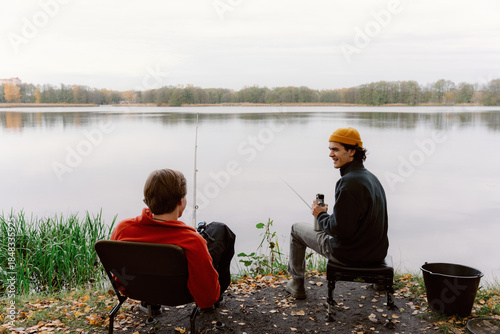 Two friends sit by a peaceful lake, fishing and sharing laughs. The autumn leaves create a colorful backdrop as they enjoy the serene atmosphere and each other's company.