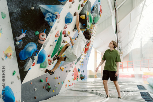 Two friends enjoy an exciting day at an indoor climbing gym. One is mid-climb on the colorful wall while the other provides encouragement from below, creating a fun atmosphere.